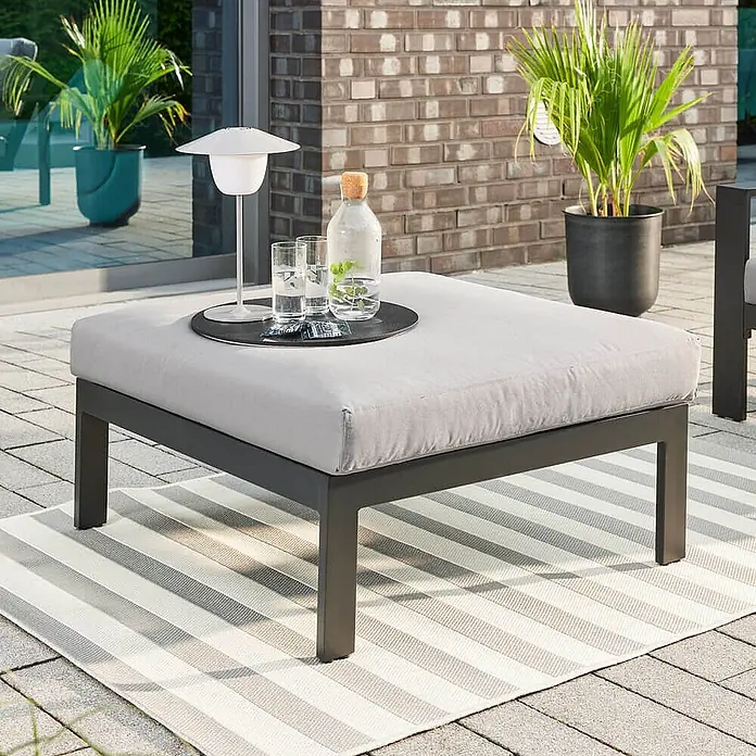 Home Deluxe Gartenhocker RIO XS -  - Dunkelgrauer Outdoor-Hocker mit hellgrauem Kissen auf einem gestreiften Teppich, als Tisch für Getränke auf einer Terrasse genutzt.