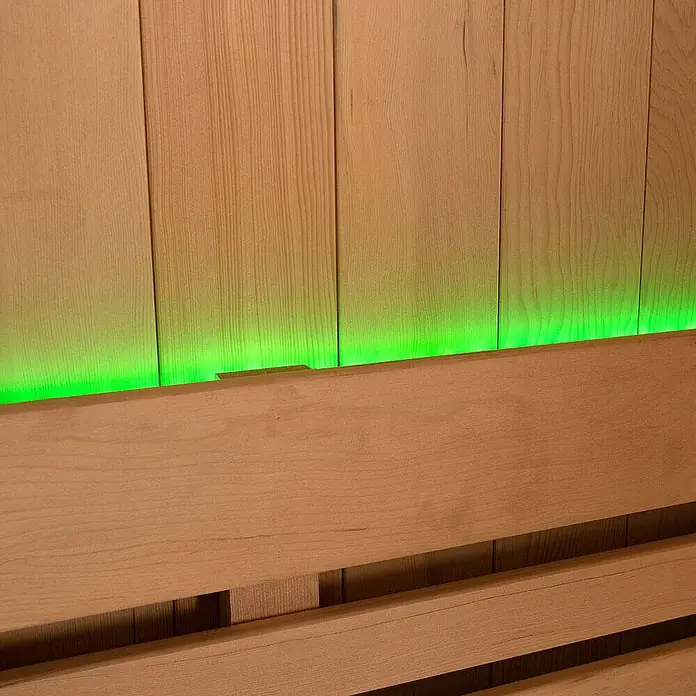 Home Deluxe Elementsauna OMAHA - Elektroofen, Steuerung Ofen: Integriert, L x B x H: 150 x 180 x 200 cm - Helle Holzwandpaneele, beleuchtet von einem grünen LED-Lichtstreifen hinter einer Holzlehne.
