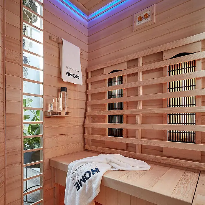 Home Deluxe Infrarotkabine REDSUN DELUXE XXL -  - Infrarot-Sauna aus hellem Holz mit sichtbaren Heizelementen, blauer LED-Beleuchtung, einer Bank und einem weißen Handtuch.