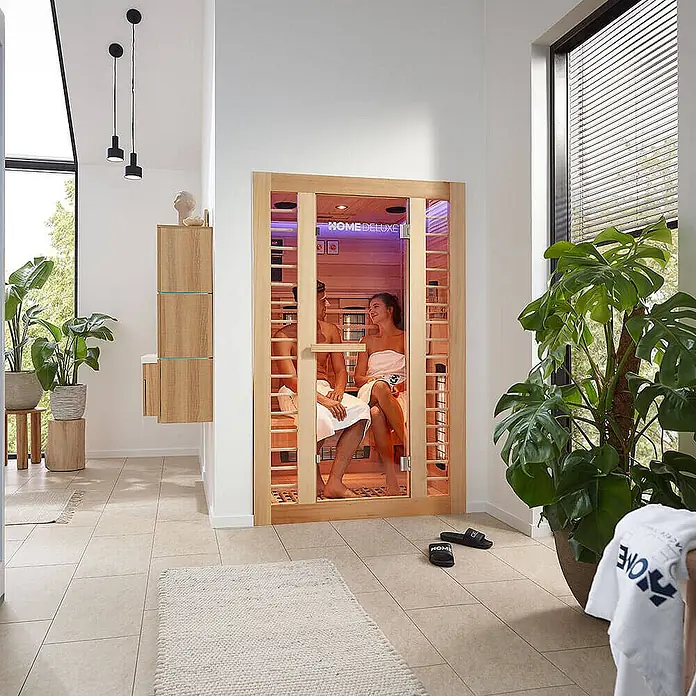 Home Deluxe Infrarotkabine REDSUM DELUXE PLUS M -  - Eine kompakte Infrarotsauna aus Holz mit Glastür, in der zwei Personen in einem modernen Wellnessbereich entspannen.