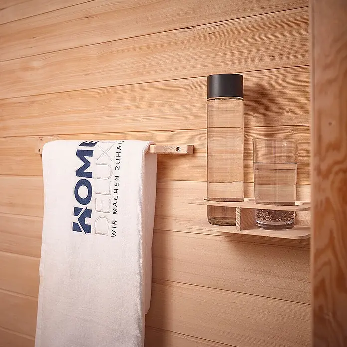 Home Deluxe Infrarotkabine REDSUM DELUXE PLUS M -  - Helle Handtuchstange aus Holz mit weißem Handtuch und passender Ablage mit Wasserflasche und Glas, an einer Saunawand.