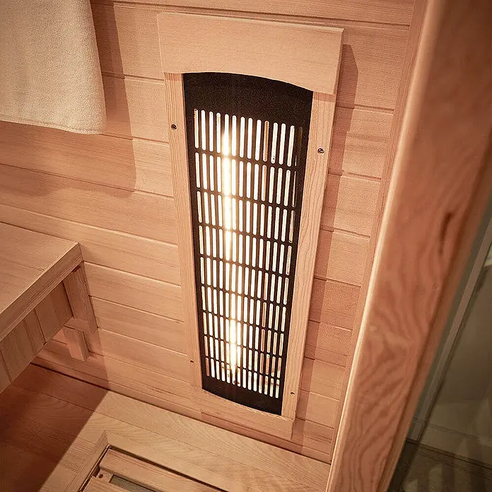 Home Deluxe Infrarotkabine REDSUN DELUXE M -  - Leuchtender Infrarotstrahler mit schwarzem Gitter an hellen Holzwänden in einer Sauna, mit Holzbank und Handtuch.