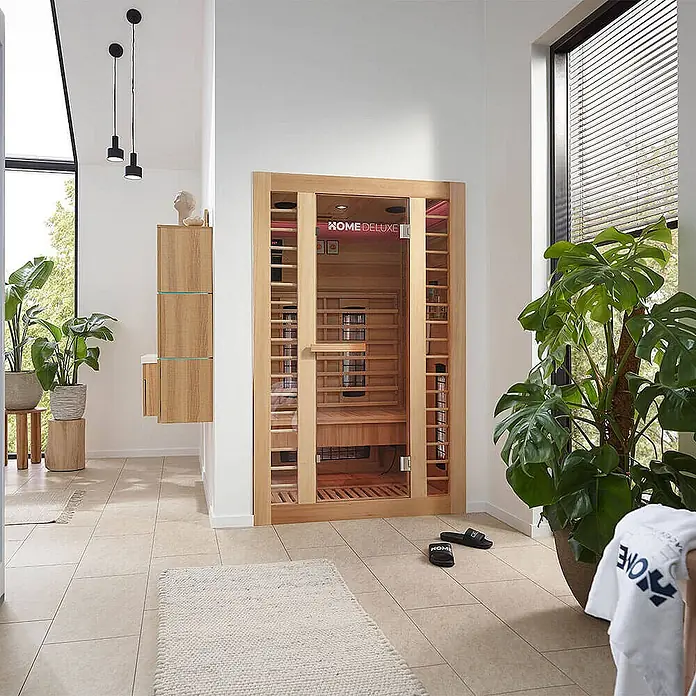 Home Deluxe Infrarotkabine REDSUN DELUXE M -  - In einem hellen, modernen Wellnessbereich steht eine Infrarotsauna aus hellem Holz mit Glastür.