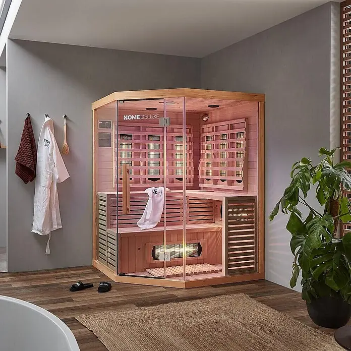 Home Deluxe Infrarotkabine SAHARA XXL -  - Eine moderne Infrarotsauna in Eckausführung aus hellem Holz und Glas, innen rosa beleuchtet, in einem Heim-Wellnessbereich.
