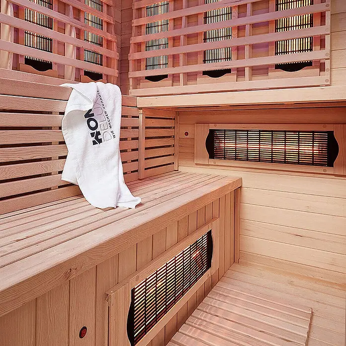 Home Deluxe Infrarotkabine SAHARA XXL -  - Innenansicht einer hellen Infrarotsauna aus Holz mit leuchtenden Heizstrahlern und weißem Handtuch auf einer Bank.