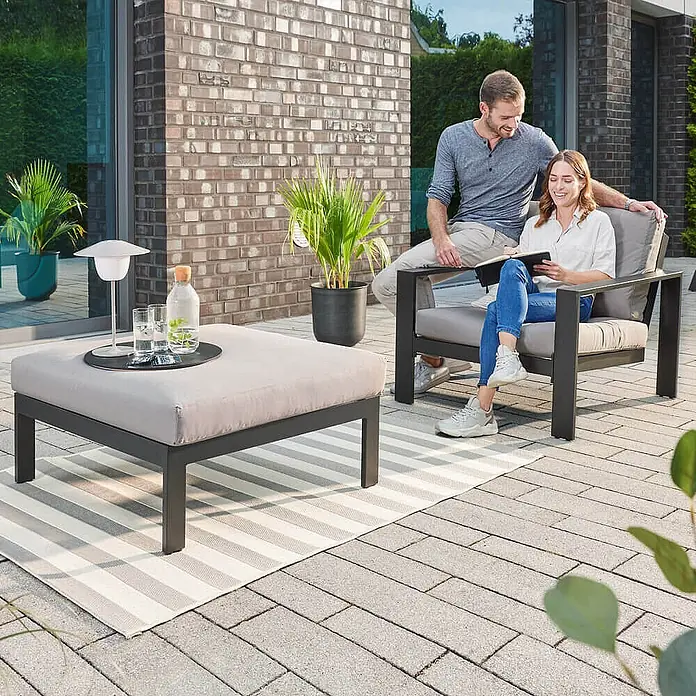 Home Deluxe Garten-Loungesessel RIO S -  - Ein Paar entspannt auf einem modernen dunkelgrauen Outdoor-Sessel und passendem Hocker mit hellgrauen Kissen auf einer Terrasse.