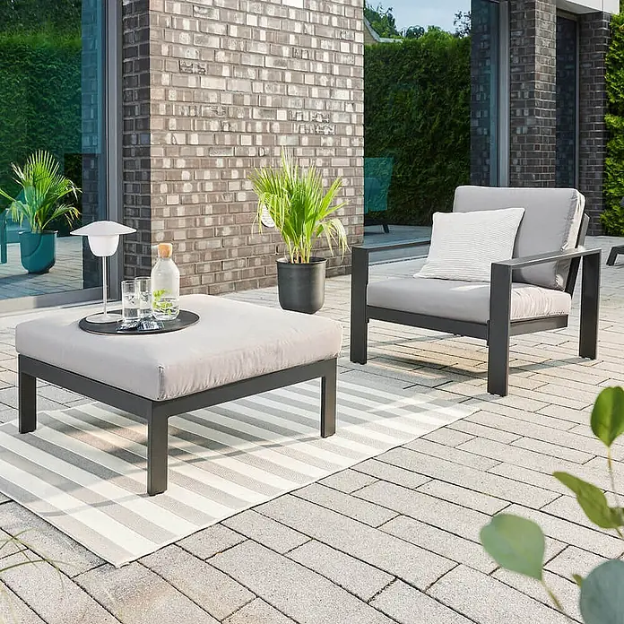 Home Deluxe Garten-Loungesessel RIO S -  - Graue Outdoor-Lounge-Sitzgruppe: Couchtisch/Hocker und Sessel mit dunklem Metallgestell und hellgrauen Kissen, auf einem Terrassenteppich.
