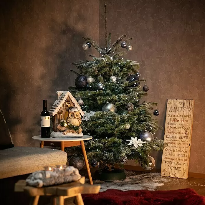 Piardino Nordmanntanne - 150 - 175 cm, Abies nordmanniana - Ein verschneiter Weihnachtsbaum, geschmückt mit silbernen und blauen Kugeln und weißen Lichtern, schafft eine festliche Winterstimmung in einem gemütlichen Raum.