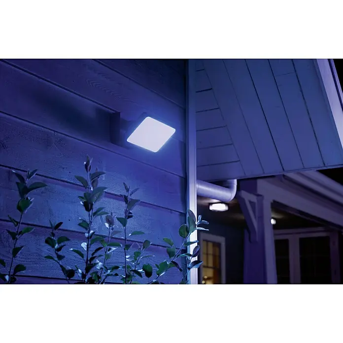 Philips Hue LED-Strahler White & Color Ambiance - 15 W, L x B x H: 22 x 16 x 15,3 cm, Schwarz, RGBW - Eine quadratische Außenleuchte spendet helles Licht neben einem Haus mit Pflanzen im Vordergrund.