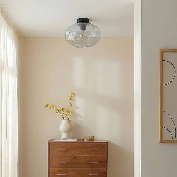 Lindby Deckenleuchte Lelia - alu / grau / zink, E27 - Transparente, organisch geformte Glas-Deckenleuchte mit schwarzem Sockel und sichtbarer Glühbirne, in einem Wohnraum installiert.