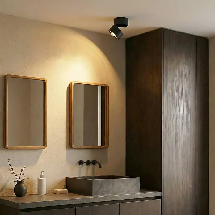 Lindby Deckenstrahler Thorid - schwarz, schwarz, G9 - Eine schwarze, verstellbare Deckenleuchte mit zwei Köpfen wirft warmes Licht auf eine strukturierte Badezimmerwand über einem Waschtisch und Spiegeln.