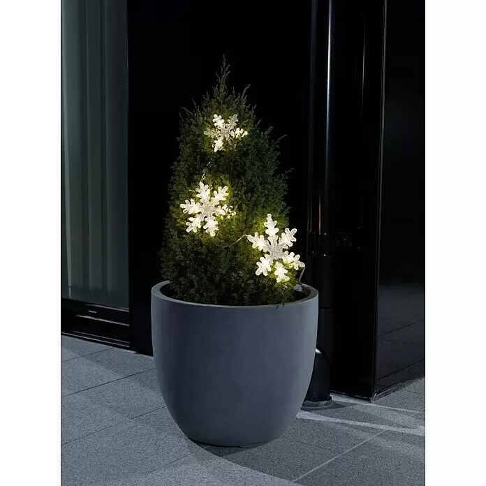 Konstsmide LED-Lichtervorhang Schneeflocken - Außen, 3,67 m, Warmweiß, 15 Stk., Batteriebetrieben - Ein Nadelbaum im Topf, verziert mit leuchtenden Schneeflocken-Lichterketten für die Außendekoration bei Nacht.