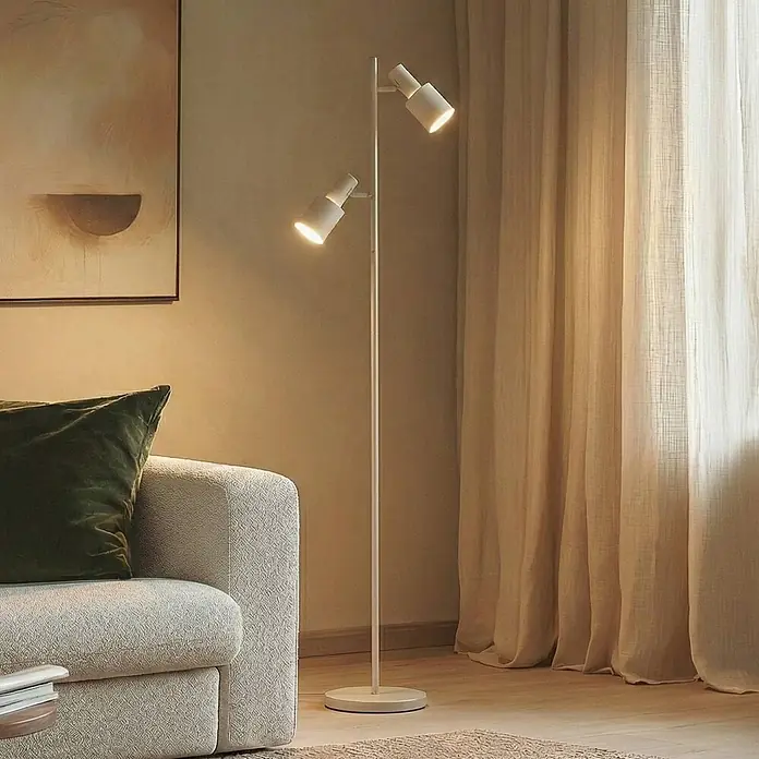Lindby Stehleuchte Ovelia - Höhe: 155 cm, creme / amber, creme / amber, E27 - Weiße Metall-Stehleuchte mit zwei verstellbaren Strahlern, beleuchtet ein Wohnzimmer neben einem hellen Sofa.
