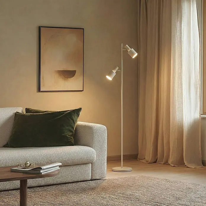 Lindby Stehleuchte Ovelia - Höhe: 155 cm, creme / amber, creme / amber, E27 - Weiße Stehlampe mit zwei verstellbaren Strahlern, die eine Wohnzimmerecke neben Sofa und Fenster beleuchtet.