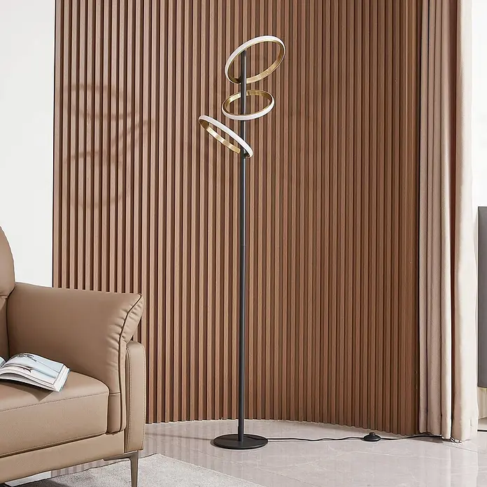Lucande Stehleuchte Madu - Höhe: 160 cm, schwarz, messing / gold - Eine moderne schwarze Stehlampe mit drei gold-weißen kreisförmigen LED-Leuchten, platziert in einem Wohnzimmer neben einer Lamellenwand.