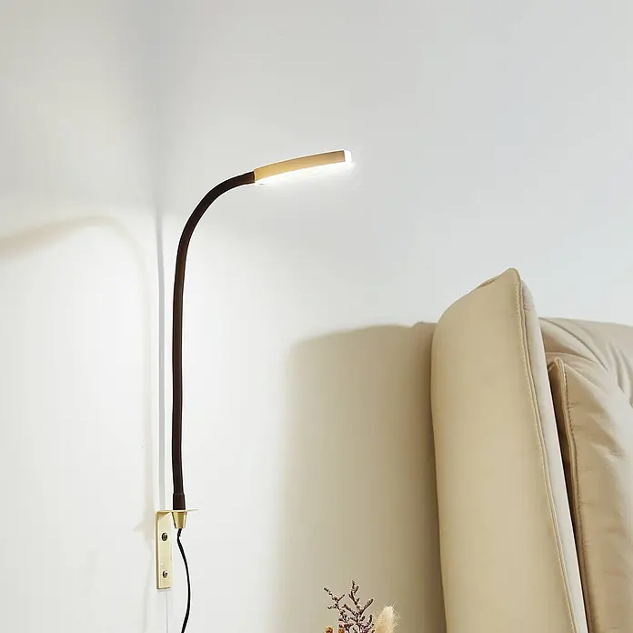 Lindby Wandleuchte Flexola - schwarz, messing / gold - Wandmontierte LED-Schwanenhalslampe mit dunklem Flexarm und Messingsockel, leuchtet neben einem cremefarbenen Sofa.