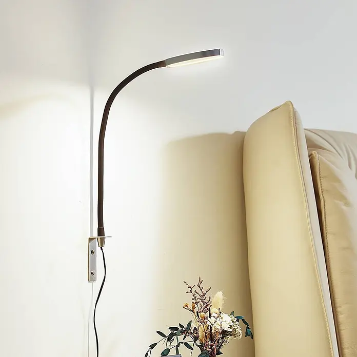 Lindby Wandleuchte Flexola - schwarz, alu / grau / zink - Wandmontierte, flexible LED-Leselampe mit dunklem Arm und Metallkopf, leuchtet neben einem beigen Sofa.