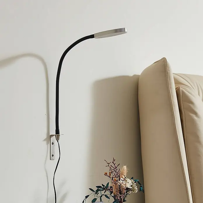 Lindby Wandleuchte Flexola - schwarz, alu / grau / zink - Wandmontierte flexible LED-Leselampe mit schwarzem Schwanenhals und silbernem Kopf, neben einem cremefarbenen Polsterkopfteil.