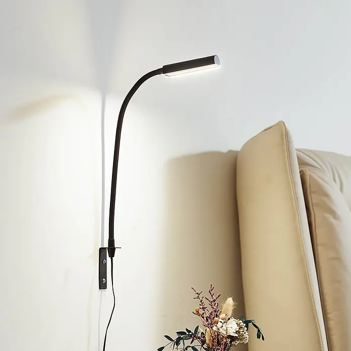 Lindby Wandleuchte Flexola - schwarz, schwarz - Schwarze wandmontierte LED-Lampe mit flexiblem Arm und linearem Leuchtkopf, spendet warmes Licht neben einem beigen Sofa.