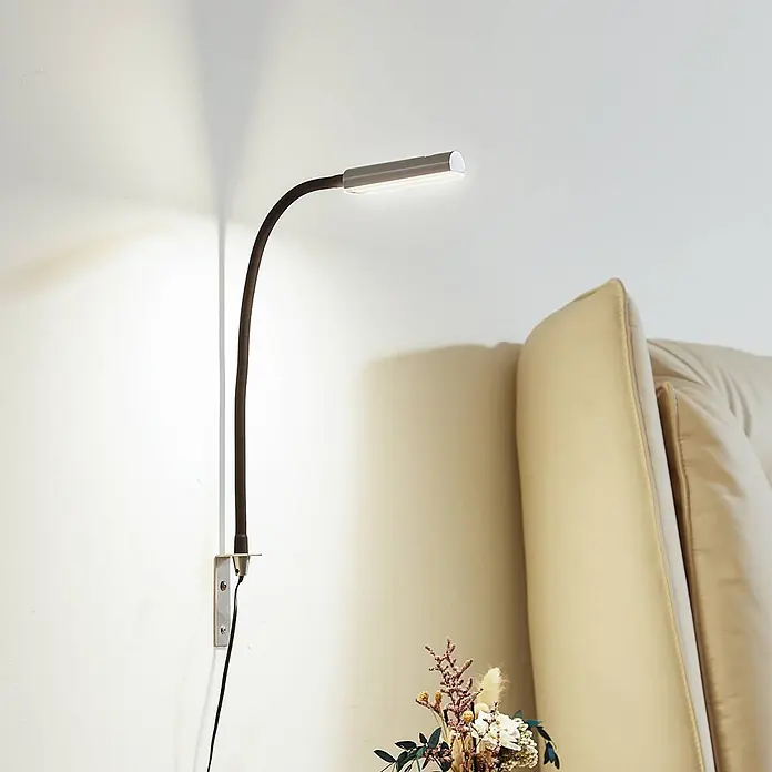 Lindby Wandleuchte Flexola - schwarz, alu / grau / zink - Wandmontierte flexible LED-Leuchte mit schwarzem Arm und silbernem Kopf, beleuchtet die Wand neben einem beigefarbenen Sofa.