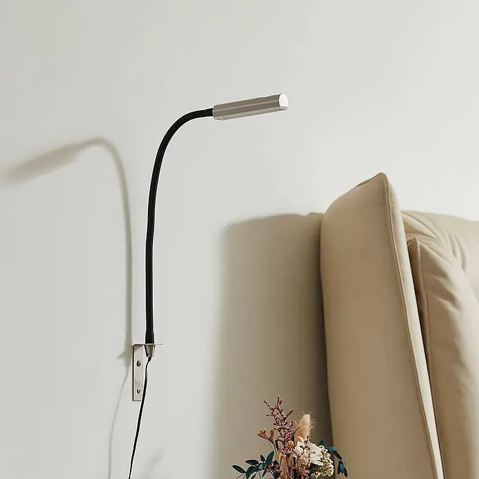 Lindby Wandleuchte Flexola - schwarz, alu / grau / zink - Wandleselampe mit schwarzem biegsamem Arm und silbernem rechteckigem Leuchtenkopf, neben einem beigem Sofa platziert.