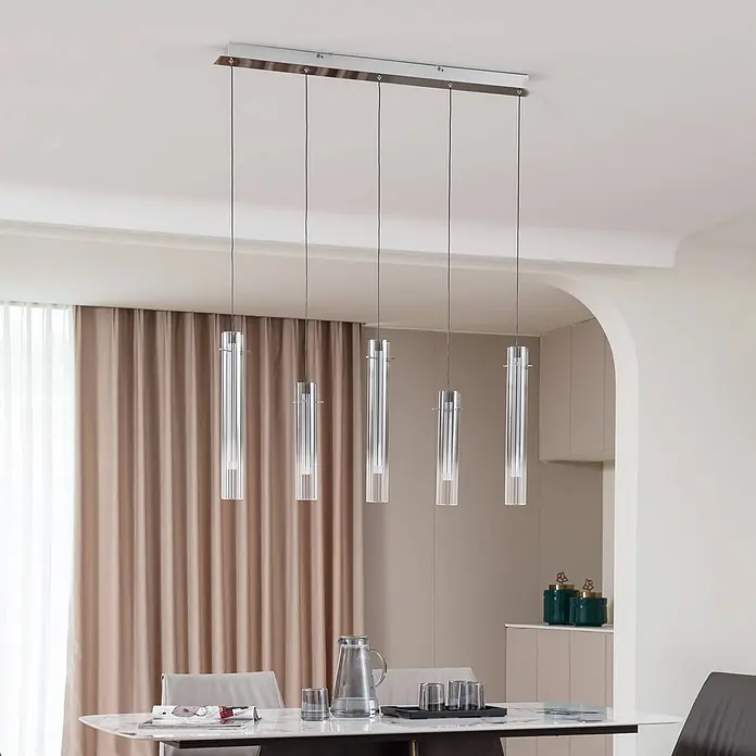 Lucande Pendelleuchte Korvitha - L x B x H: 92 x 8,5 x 45 cm, alu / grau / zink, rauchgrau, silber - Fünf zylindrische Pendelleuchten aus klarem Glas mit rechteckigem Chromsockel hängen über einem Esstisch.