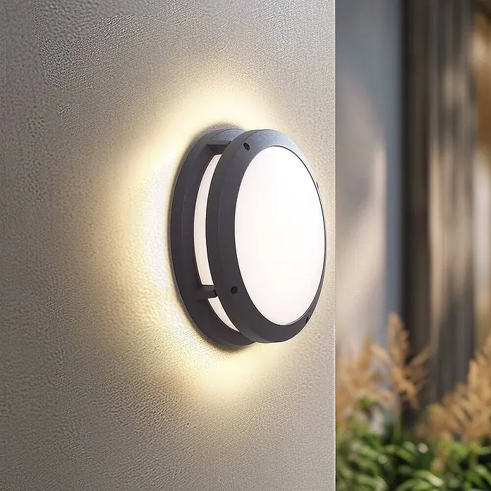 Lindby Außenwandleuchte Liren - alu / grau / zink, IP54 - Dunkelgraue, runde Außenwandleuchte strahlt warmes Licht auf eine strukturierte Wand.