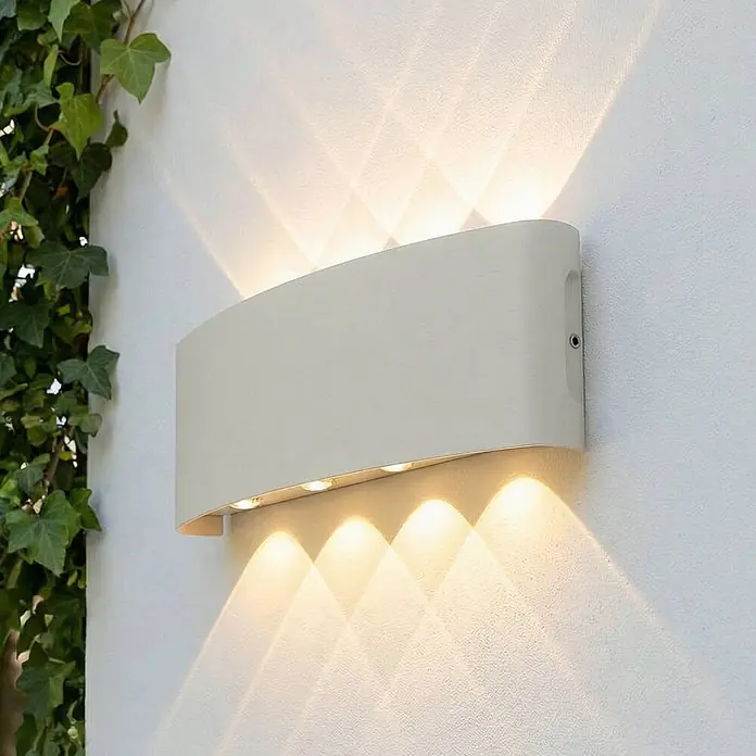 Lindby Außenwandleuchte Ithan - weiß / opal, IP54 - Eine cremefarbene, gebogene Außenwandleuchte beleuchtet eine strukturierte Wand nach oben und unten, daneben rankt Efeu.
