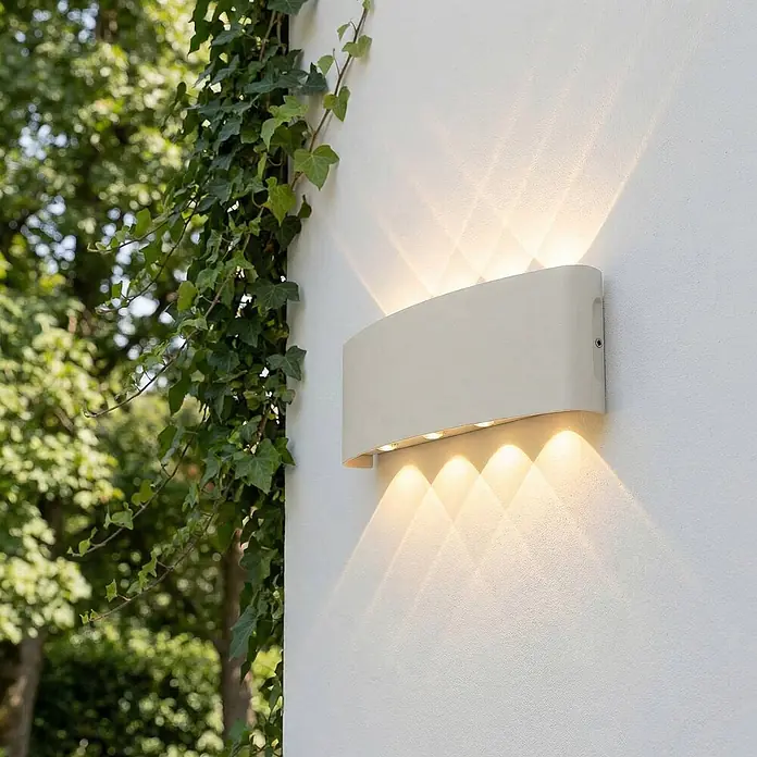 Lindby Außenwandleuchte Ithan - weiß / opal, IP54 - Gekrümmte, cremefarbene Außenwandleuchte an einer Wand neben Efeu, die nach oben und unten gemustertes Licht wirft.