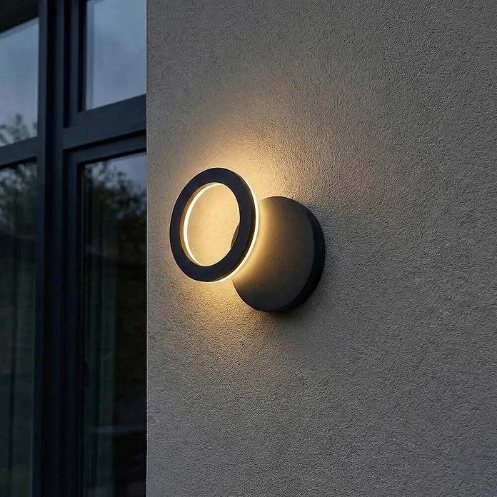 Lindby Außenwandleuchte Ilveo - schwarz, schwarz, IP54 - Dunkle runde Außenwandleuchte an einer strukturierten Wand, die warmes Licht abstrahlt, neben einem Fenster.