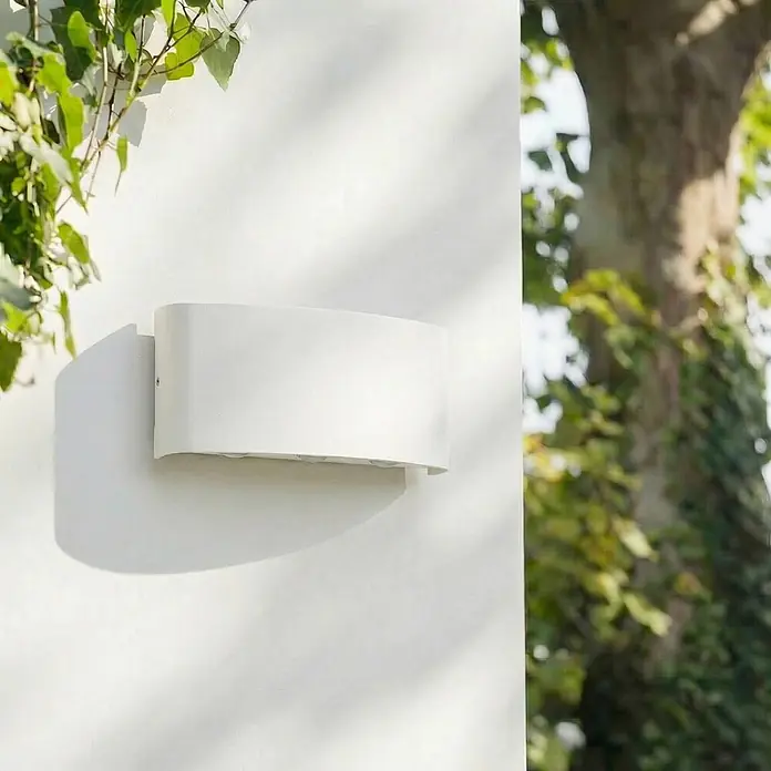 Lindby Außenwandleuchte Ithan - weiß / opal, IP54 - Weiße Außenwandleuchte an einer hellen Wand, umgeben von grünem Laub.