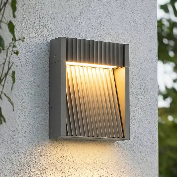Lucande Außenwandleuchte Tavion - schwarz, schwarz, IP65 - Graue, quadratische Außenwandleuchte mit geripptem Innenbereich, die warmes Licht nach oben und unten auf eine strukturierte Wand wirft.