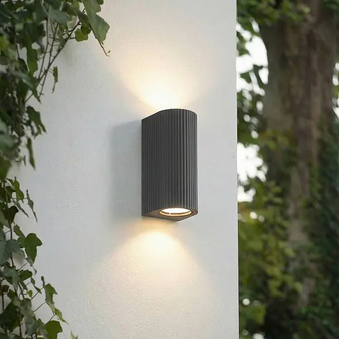 Lindby Wandleuchte aussen in schwarz, Aussenlampe Wand -  - Dunkelgraue geriffelte Außenwandleuchte, die warmes Licht nach oben und unten auf eine strukturierte Wand wirft.