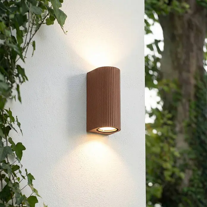 Lindby Wandleuchte aussen in rost, Aussenlampe Wand -  - Braune gerippte Außenwandleuchte, die warmes Licht nach oben und unten auf eine strukturierte weiße Wand wirft.