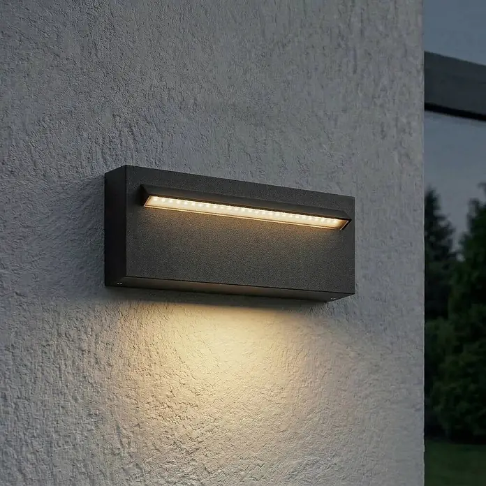 Lucande Außenwandleuchte Joril - schwarz, schwarz, IP65 - Dunkelgraue rechteckige LED-Wandleuchte beleuchtet eine strukturierte Fläche mit warmweißem Licht.