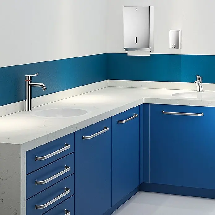 Delabie Spültischarmatur Delabie -  - Edelstahl-Handtuchspender und Seifenspender über weißen Waschbecken mit Chromarmaturen und blauen Schränken in modernem Waschraum.