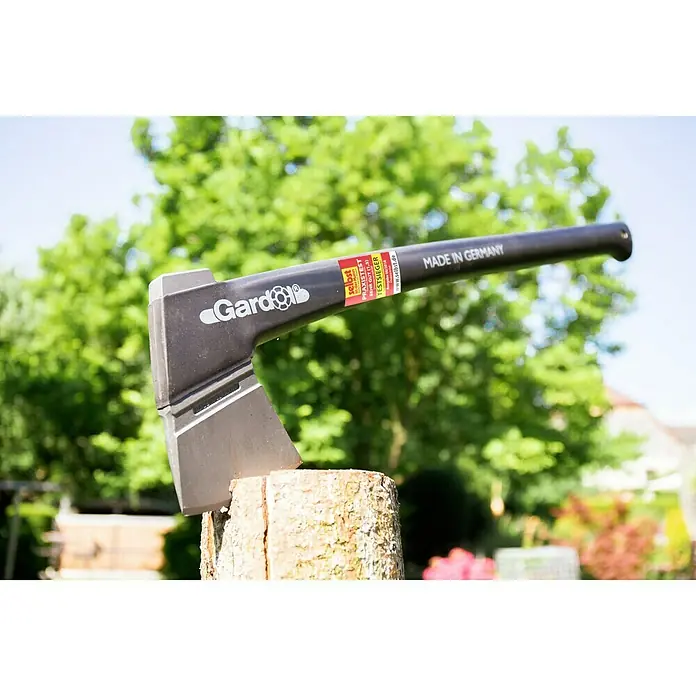 Gardol Profilinie Universalbeil - Länge: 37 cm - Axt von Gardol steckt in einem Holzblock im Freien vor Bäumen und einem Haus.