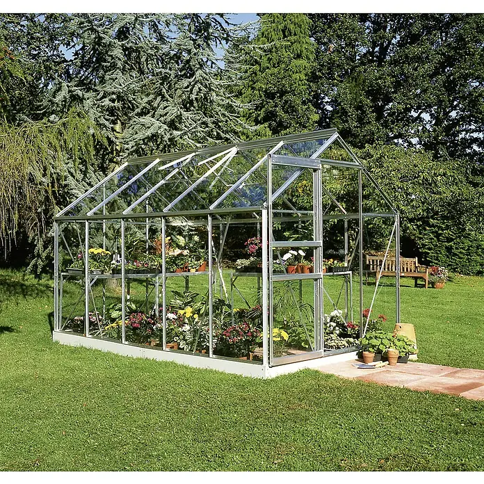 Halls Gewächshaus Popular 106 - Außenmaß inkl. Dachüberstand (B x T): 193 x 319 cm, Silber, Wandstärke: 3 mm - Das Bild zeigt ein Gewächshaus mit Glaswänden, gefüllt mit bunten Blumen auf Regalen, in einem grünen Garten.