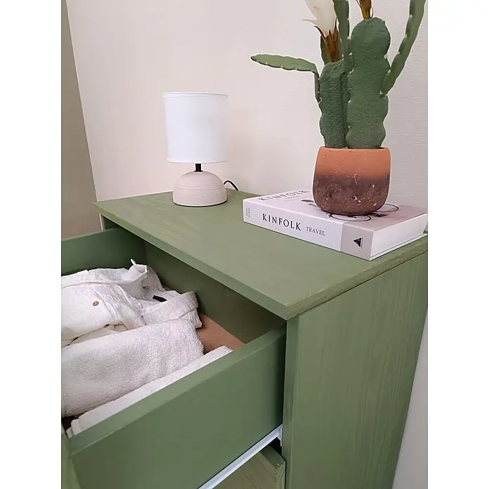 Astigarraga Vita Cómoda - L x An x Al: 36 x 65 x 96 cm, Natural, Número de cajones: 4 ud. - Cómoda de madera verde salvia con un cajón abierto que revela ropa y toallas blancas. Encima hay una lámpara, un libro y un cactus en maceta.