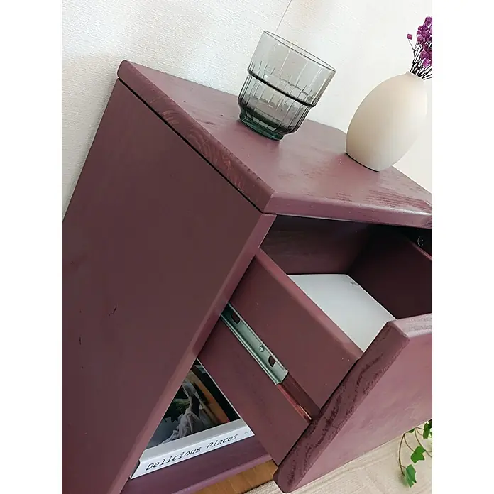 Astigarraga Vita Mesita de noche - L x An x Al: 25 x 40 x 50 cm, Natural, Número de cajones: 1 ud. - Mesita de noche de madera morado oscuro con cajón y estante abiertos. Vaso y jarrón arriba, libro en el estante.