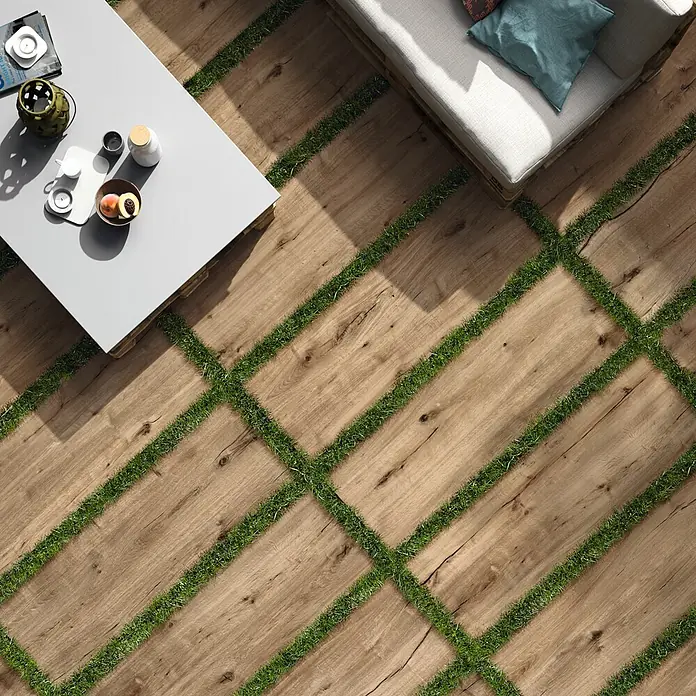 Terrassenfliese Essenze 2.0 - 40 x 120 x 2 cm, Honey, Matt - Outdoor-Terrassenboden aus hellen Holzoptik-Dielen mit grünen Grasstreifen, mit Tisch und Sofa.