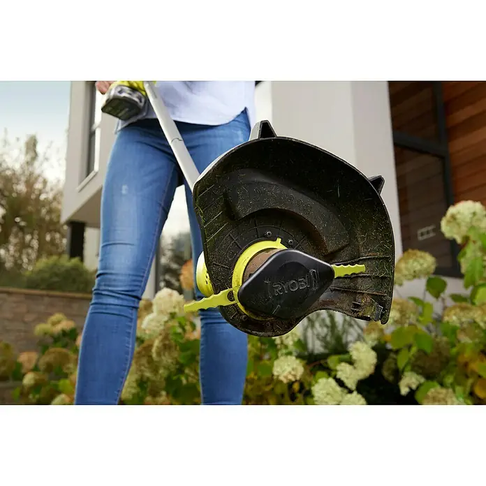 Ryobi Kunststoffmesser RAC155 - Passend für: Ryobi Akku-Rasentrimmer mit ‚Auto-Feed-Funktion‘ - Nahaufnahme eines Ryobi Rasentrimmers mit Schutzvorrichtung und Schneidmesser vor einem Garten mit blühenden Büschen.