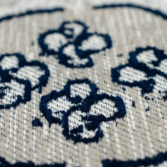 Kayoom Flachgewebeteppich River 120 - Grau/Schwarz, 170 x 120 cm, 70 % Baumwolle, 30 % Polyester - Nahaufnahme eines Stoffmusters mit stilisierten blauen Blüten auf beigem Grund, ideal für Dekorationen und Textilien.