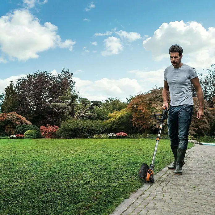 Worx PowerShare 20V Akku-Rasentrimmer WG163E.9 - 20 V, Li-Ionen, Ohne Akku - Ein Mann benutzt einen Worx Rasenkantenschneider auf einem gepflasterten Weg, um Rasenkanten zu trimmen. Der Trimmer ist orange und silber.