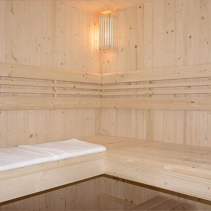 Home Deluxe Elementsauna SHADOW BIG XL - Elektroofen, Steuerung Ofen: Integriert, L x B x H: 200 x 200 x 190 cm - Sauna-Innenraum mit hellen Holzwänden, Bänken, einem weißen Handtuch und einer Eckwandleuchte.