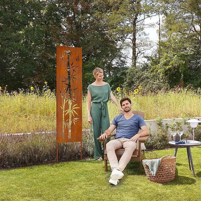 Home Deluxe Sichtschutzelement AURELIA - Stahl, Orange - Gartensichtschutz aus Cortenstahl mit Bambusmotiv, in einer üppigen Gartenszene mit Personen.