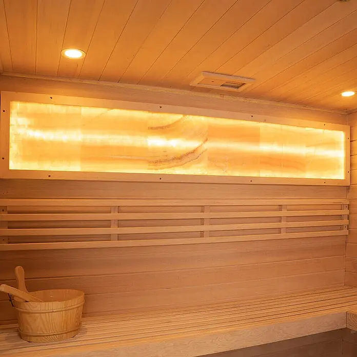 Home Deluxe Elementsauna NEOLA - Elektroofen, Steuerung Ofen: Integriert - Hölzerne Saunainterieur mit leuchtendem, hinterleuchtetem Onyx-Paneel über Lattenbänken und einem Holzeimer mit Schöpfkelle.