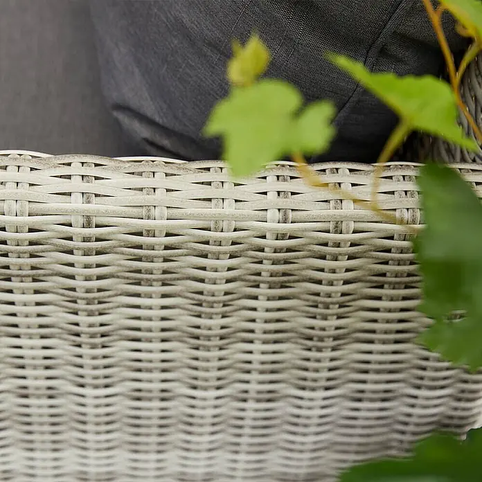 Home Deluxe Polyrattan Sitzgruppe BAHIA Mit Regenabdeckung -  - Nahaufnahme von hellgrauen, geflochtenen Gartenmöbeln mit dunkelgrauem Kissen und grünen Blättern.