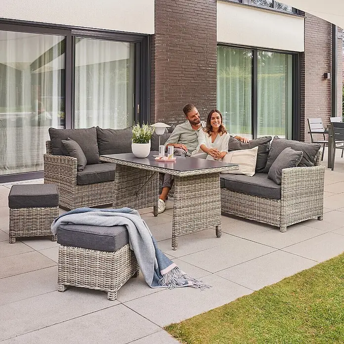 Home Deluxe Polyrattan Sitzgruppe BAHIA Mit Regenabdeckung -  - Helles Rattan-Gartenlounge-Set mit dunkelgrauen Kissen und einem Tisch, von einem Paar auf einer Terrasse genutzt.