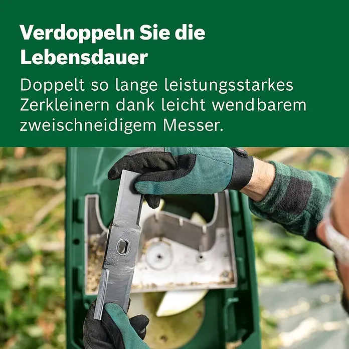 Bosch Elektro-Messerhäcksler Axt Rapid 2000 - Max. Aststärke: 3,5 cm, 2.000 W - Behandschuhte Hände halten ein umkehrbares zweischneidiges Metallmesser über einem geöffneten grünen Gartenhäcksler.